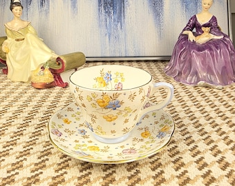 May include: A vintage teacup and saucer set with a floral pattern in yellow, blue, and pink. The teacup has a light blue handle and rim. Two porcelain figurines are in the background, along with an abstract painting.