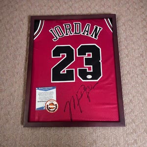 May include: Framed red basketball jersey with the name "JORDAN" and the number "23" in black and white. The jersey is signed and includes a certification sticker and a Beckett Certificate.