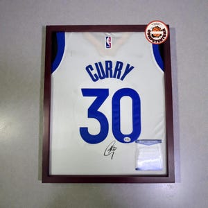 May include: Framed white basketball jersey with blue lettering and the number 30. The jersey is signed and includes a certificate of authenticity. The frame is dark brown.