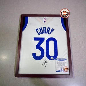 May include: Framed white basketball jersey with the name "CURRY" and the number "30" in blue. The jersey is signed and includes a Beckett Certificate of Authenticity. The frame is a dark brown color.