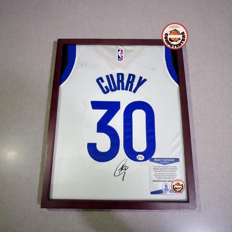 May include: Framed white basketball jersey with the name "CURRY" and the number "30" in blue. The jersey is signed and includes a Beckett Certificate of Authenticity. The frame is a dark brown color.
