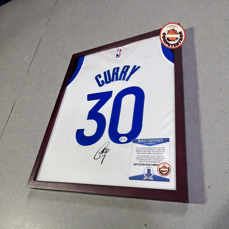 May include: Framed white basketball jersey with blue accents, displaying the name "CURRY" and the number "30" in blue. Includes an autograph and a Beckett Certificate of Authenticity. The jersey is encased in a dark brown frame.