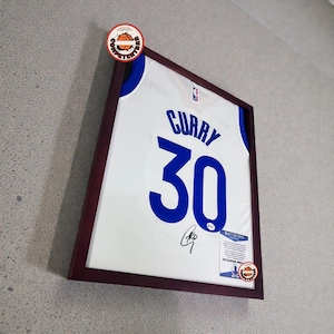 May include: Framed white basketball jersey with blue lettering and the number 30. The jersey is signed and displayed in a dark wood frame. A basketball-themed sticker is affixed to the frame.