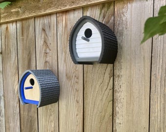 May include: Two birdhouses are mounted on a wooden fence. The birdhouses have a unique design with a dark gray shingled roof and a white or light wood front. One birdhouse has a blue trim.