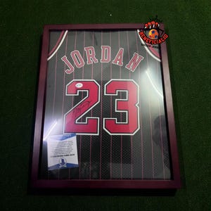 May include: Framed basketball jersey with the name "JORDAN" and the number "23" in red on a black jersey. The jersey is signed and includes a certificate of authenticity. The frame is a dark brown color.