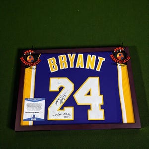 Op de afbeelding: Ingelijst paars en goudkleurig shirt met de naam "BRYANT" en het nummer "24". Het shirt is gesigneerd en bevat een Beckett-certificaat. De lijst heeft decoratieve voetbalaccenten.