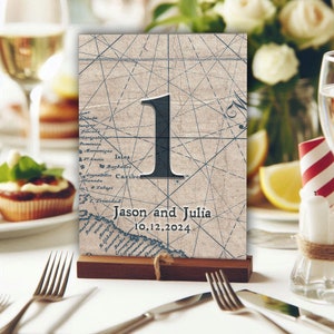 May include: A vintage-style table number card with a nautical map background. The number "1" is printed in black, and the text "Jason and Julia 10.12.2024" is printed in black at the bottom of the card.