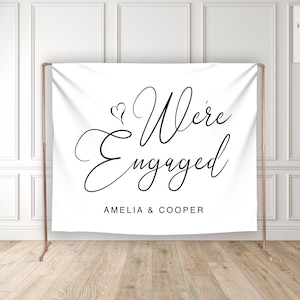May include: White backdrop with black calligraphy that reads "We're Engaged" with the names Amelia & Cooper below.