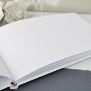 May include: A white blank guest book with a white cover and white pages. The book is open and lying flat on a gray surface.