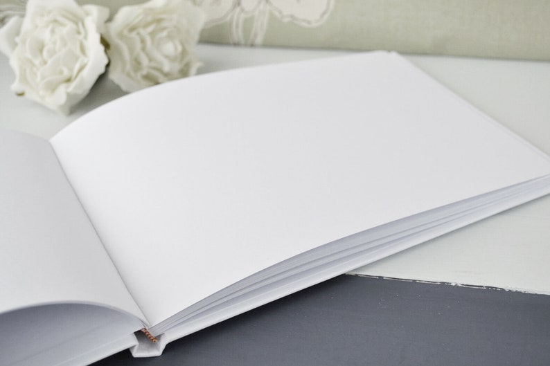 May include: A white blank guest book with a white cover and white pages. The book is open and lying flat on a gray surface.