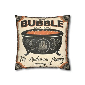 May include: A cream-colored pillow with a black and orange Halloween-themed design. The pillow features a cauldron filled with orange pumpkins and the text "Bubble Toil and Trouble The Anderson Family Brewing Co."