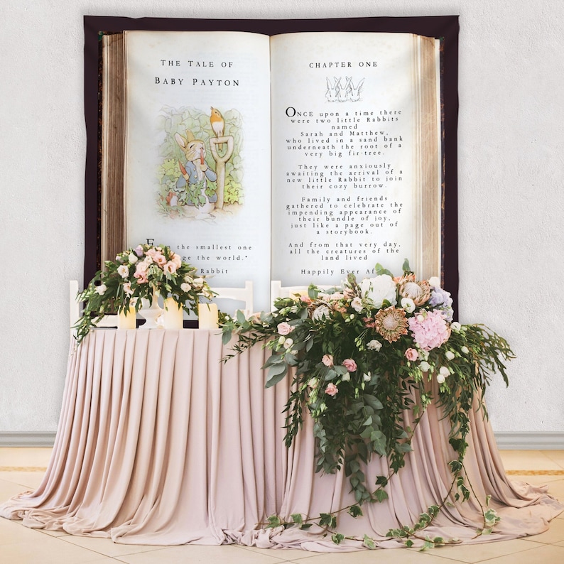May include: A wedding table decorated with a large open book backdrop, featuring illustrations and text from "The Tale of Baby Payton." The table is draped in a blush-coloured fabric and decorated with floral arrangements in pink, white, and green.