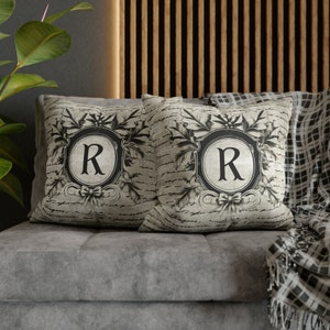 May include: Two decorative throw pillows with a vintage, distressed look. Each pillow features a black and white design with a large letter "R" in a circle surrounded by ornate floral details.