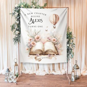 May include: A white backdrop with a watercolor illustration of a book, flowers, and a hot air balloon. The text reads "A new chapter begins Alexis turns one first birthday".