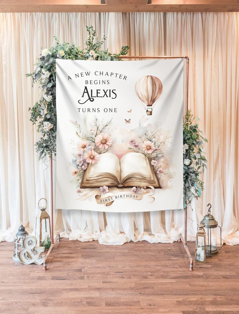 May include: A white backdrop with a watercolor illustration of a book, flowers, and a hot air balloon. The text reads "A new chapter begins Alexis turns one first birthday".