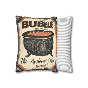 May include: A decorative pillow with a vintage-style illustration of a cauldron with orange liquid and the text "Bubble Toil and Trouble The Anderson Family Brewing It". The pillow is paired with a gray and white striped pillow.