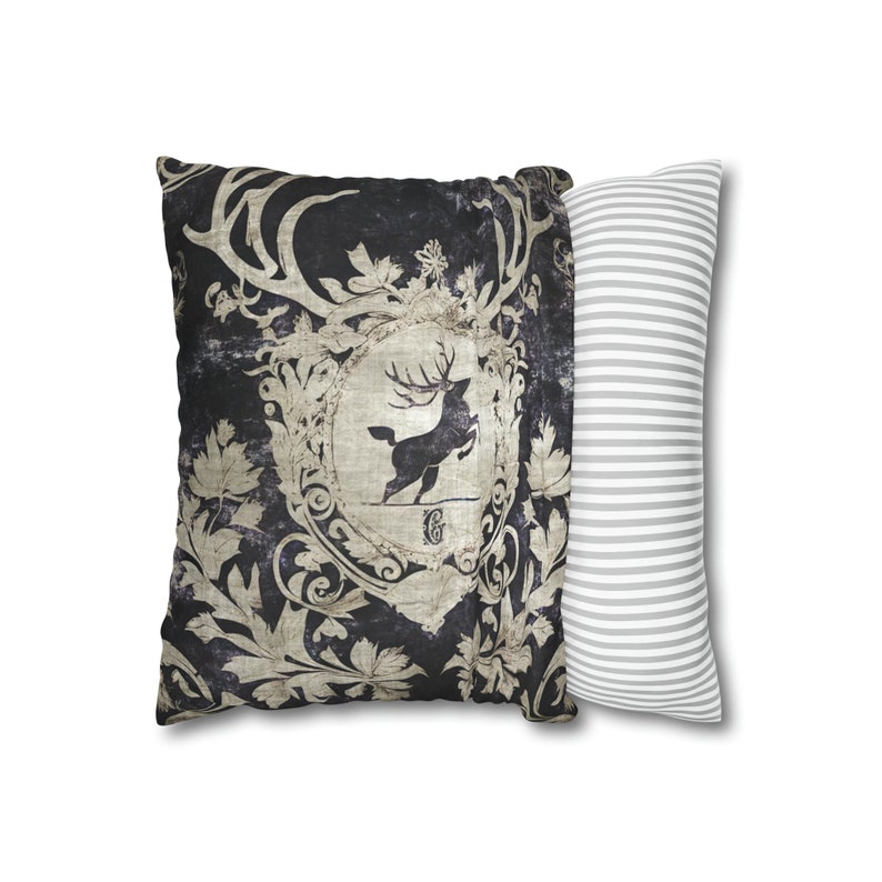 May include: A decorative throw pillow with a black background and a vintage-style design featuring a stag with antlers and ornate floral details. The pillow is paired with a white pillow with gray stripes.