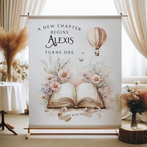 May include: A white backdrop with a watercolor illustration of an open book with flowers and a hot air balloon. The text reads "A new chapter begins Alexis turns one first birthday".