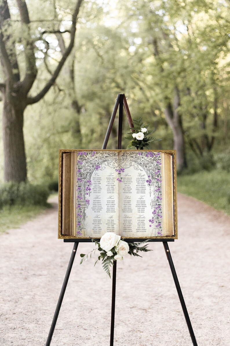 May include: A wedding seating chart displayed on an easel. The chart is printed on a vintage-style book with a gold frame. The book is open to two pages, each with a list of names and table assignments. The pages are decorated with purple flowers and a floral archway design.