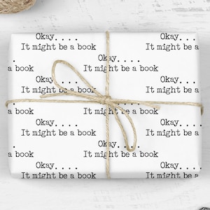 May include: White wrapping paper with the text "Okay... It might be a book" repeated multiple times. The paper is wrapped around a gift box and tied with twine. There are scissors and paper clips on a white wooden surface.