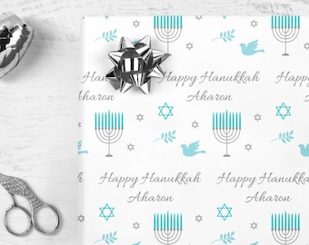 May include: White wrapping paper with a repeating pattern of blue menorahs, gray stars, gray doves, and gray branches. The text "Happy Hanukkah Aharon" is repeated throughout the pattern.
