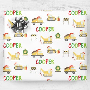 May include: White wrapping paper with a repeating pattern of yellow construction trucks wearing Santa hats, green wreaths, candy canes, and red and green "COOPER" text.