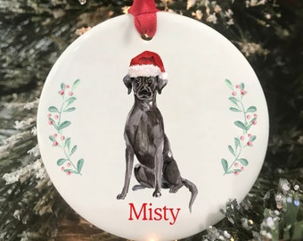 May include: A white ceramic Christmas ornament featuring a black Labrador Retriever wearing a red Santa hat. The dog is sitting and the ornament has a green wreath with red berries on either side. The name "Misty" is written in red at the bottom of the ornament.