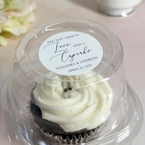 May include: A clear plastic container with a cupcake inside. The cupcake is frosted with white frosting and has a chocolate base. The container has a white sticker on it that says "All you need is Love and a Cupcake" with the names Natasha & Andrew and the date April 29, 2024.