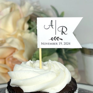 May include: A white cupcake with white frosting and a flag on top. The flag is white with black lettering that says "A|R" and "NOVEMBER 19, 2024".