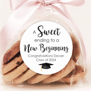 May include: A white circle sticker with black text that reads "A Sweet ending to a New Beginning Congratulations Devan Class of 2024". A black graduation cap with a gold tassel is below the text. The sticker is on a clear plastic bag filled with cookies and tied with a pink ribbon.