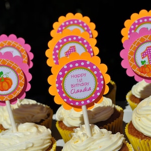 May include: Pink and orange cupcake toppers with a pumpkin design and the text "Happy 1st Birthday Claudia".
