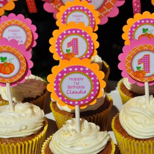 May include: A table with cupcakes decorated with orange, pink, and white frosting. The cupcakes are topped with orange, pink, and white pumpkin-themed toppers that say "Happy 1st Birthday Claudia".