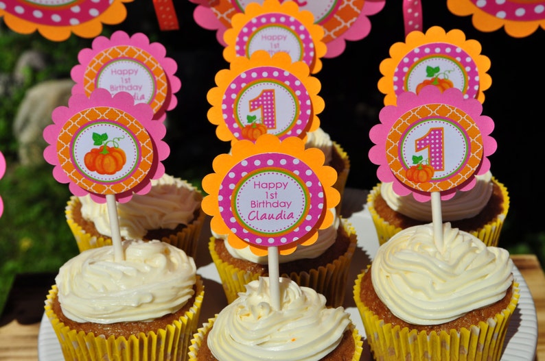 May include: A table with cupcakes decorated with orange, pink, and white frosting. The cupcakes are topped with orange, pink, and white pumpkin-themed toppers that say "Happy 1st Birthday Claudia".