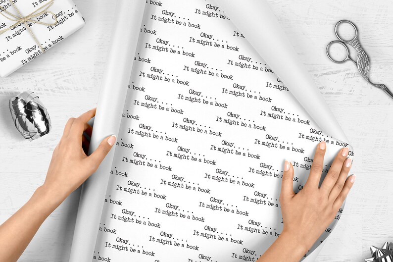 May include: White wrapping paper with the repeated text "Okay... It might be a book" printed on it. A person is holding the paper and rolling it up.