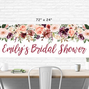 Op de afbeelding: Een witte banner met een bloemenontwerp in tinten roze, bordeauxrood en groen. De banner heeft de tekst "Emily's Bridal Shower" in bordeauxrode schrift. De banner is 183 cm breed en 61 cm hoog.