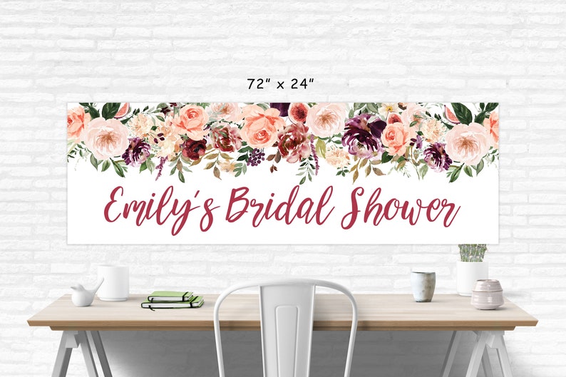 Op de afbeelding: Een witte banner met een bloemenontwerp in tinten roze, bordeauxrood en groen. De banner heeft de tekst "Emily's Bridal Shower" in bordeauxrode schrift. De banner is 183 cm breed en 61 cm hoog.