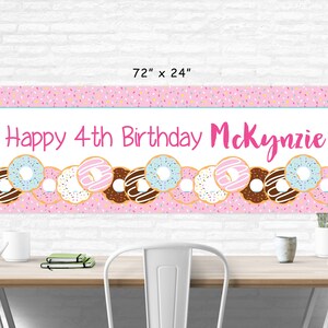 Op de afbeelding: Roze en witte donut-thema verjaardagsbord met de tekst "Happy 4th Birthday McKenzie" en een rij kleurrijke donuts met sprinkles.