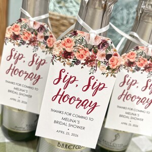 以下が含まれることがあります： 「Sip, Sip Hooray」と赤い筆記体で書かれたタグが付いたシャンパンボトル3本。タグはピンク、ピーチ、バーガンディの色合いの花柄で飾られています。ボトルは白いリボンで結ばれており、ブライダルシャワーに最適です。