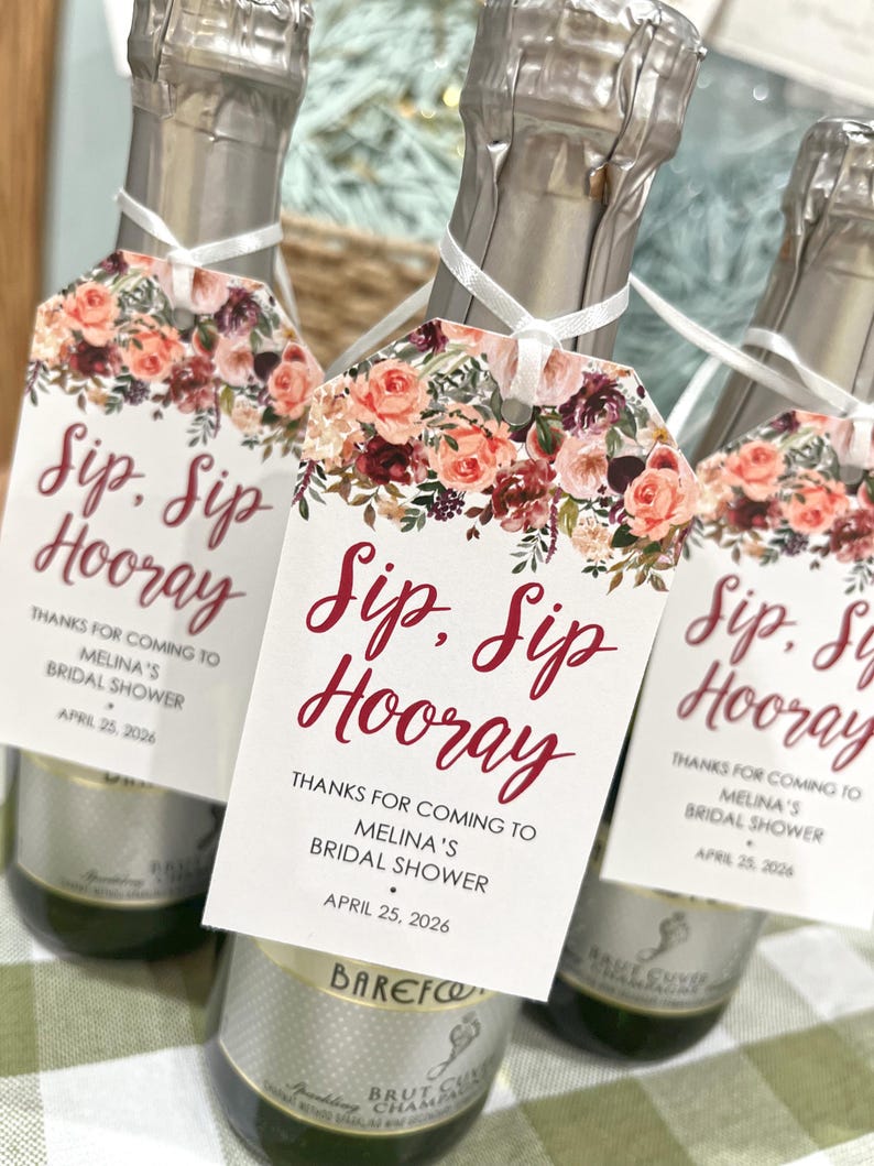以下が含まれることがあります： 「Sip, Sip Hooray」と赤い筆記体で書かれたタグが付いたシャンパンボトル3本。タグはピンク、ピーチ、バーガンディの色合いの花柄で飾られています。ボトルは白いリボンで結ばれており、ブライダルシャワーに最適です。