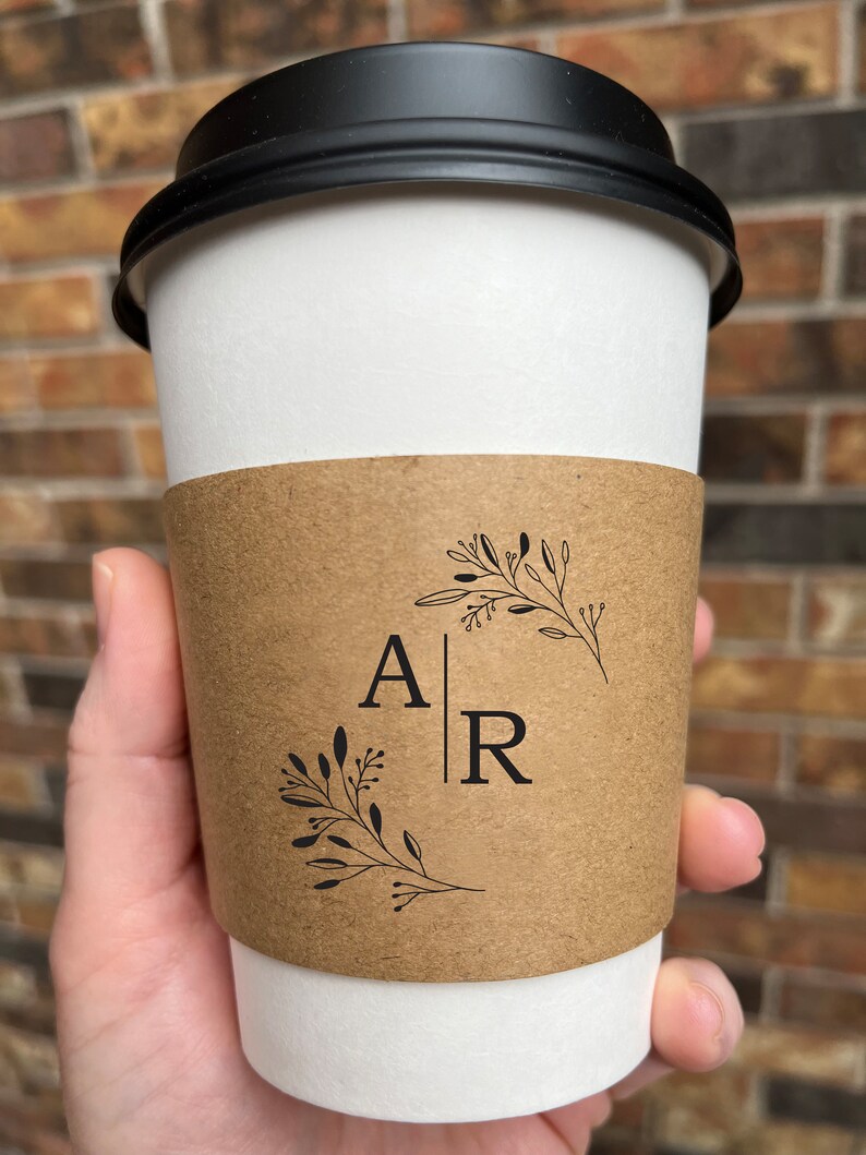 May include: A white paper coffee cup with a black lid, held by a hand. The cup has a brown sleeve with the letters "A" and "R" separated by a vertical line, and floral designs.