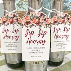 以下が含まれることがあります： 「Sip, Sip Hooray」と「Melina's Bridal Shower、2026年4月25日」と書かれたタグが付いたシャンパンボトル3本。タグはピンク、赤、バーガンディの色合いの花柄で飾られています。ブライダルシャワー用です。