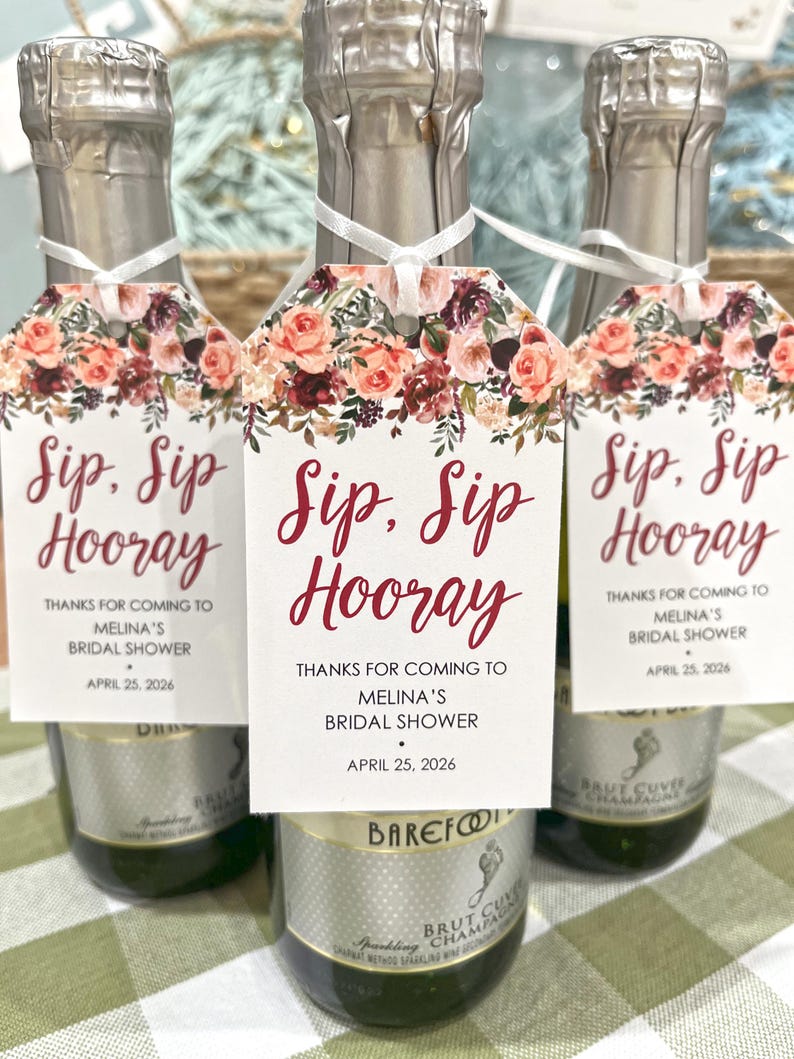 以下が含まれることがあります： 「Sip, Sip Hooray」と「Melina's Bridal Shower、2026年4月25日」と書かれたタグが付いたシャンパンボトル3本。タグはピンク、赤、バーガンディの色合いの花柄で飾られています。ブライダルシャワー用です。