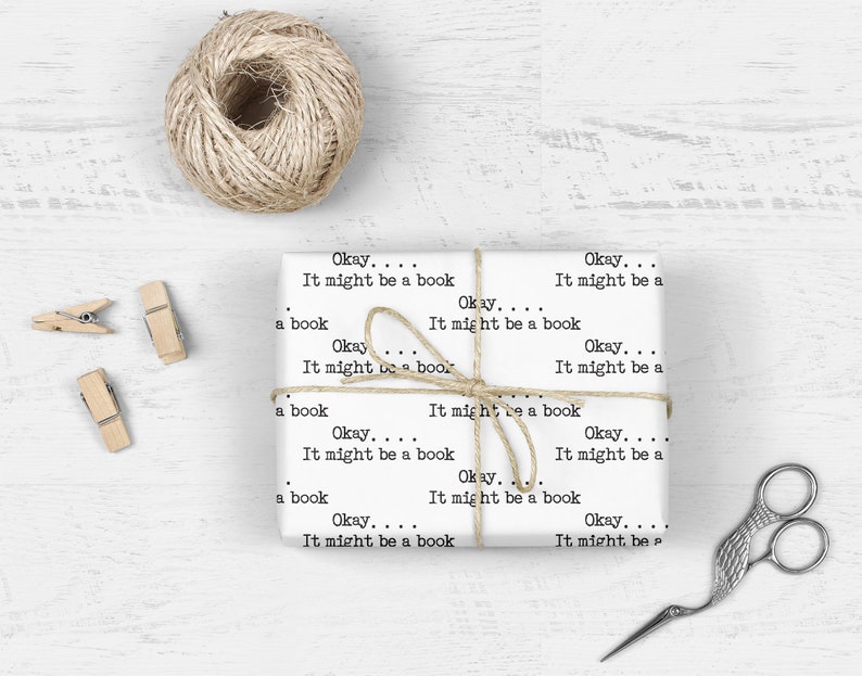 May include: White wrapping paper with the text "Okay... It might be a book" repeated multiple times. The paper is wrapped around a gift box and tied with twine. There are scissors and paper clips on a white wooden surface.