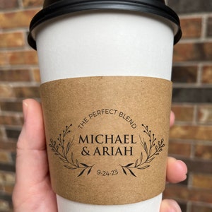 May include: A white paper coffee cup with a black lid. A brown paper sleeve wraps around the cup, featuring the text "THE PERFECT BLEND" above the names "MICHAEL & ARIAH" and the date "9-24-23", all within a floral wreath.