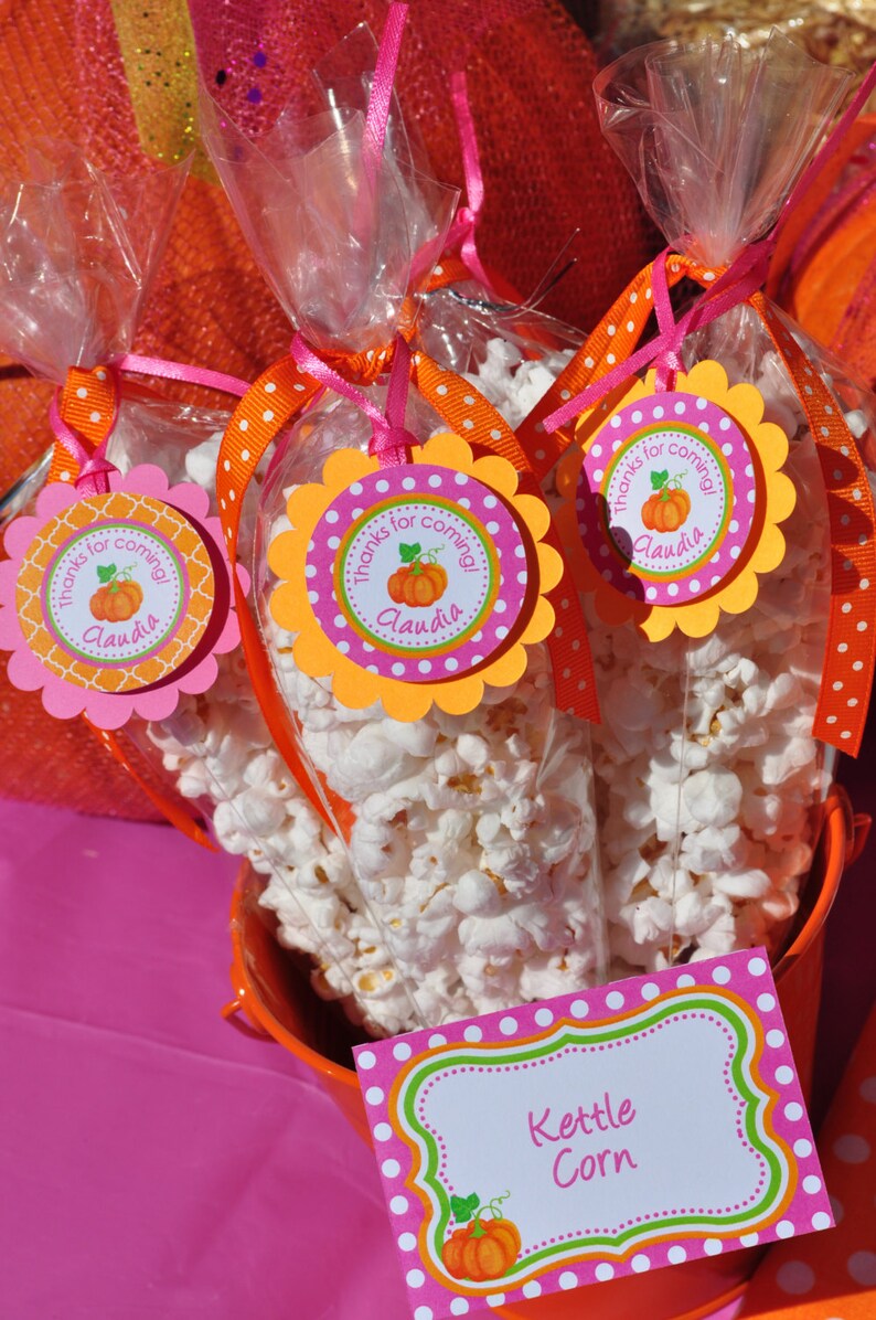 May include: A small orange bucket filled with white popcorn. Three clear bags of popcorn are tied with pink and orange ribbon and have a pink and orange pumpkin-themed tag that says "Thanks for coming! Claudia". A pink and green sign with white polka dots says "Kettle Corn".