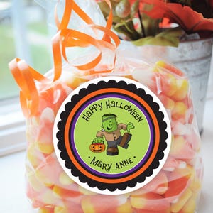 May include: A round sticker with a black scalloped border, an orange and black striped border, a purple border, and a green background. The sticker says "Happy Halloween" in black text and features a cartoon green Frankenstein monster holding a jack-o-lantern. The sticker also says ". Mary Anne ." in black text.