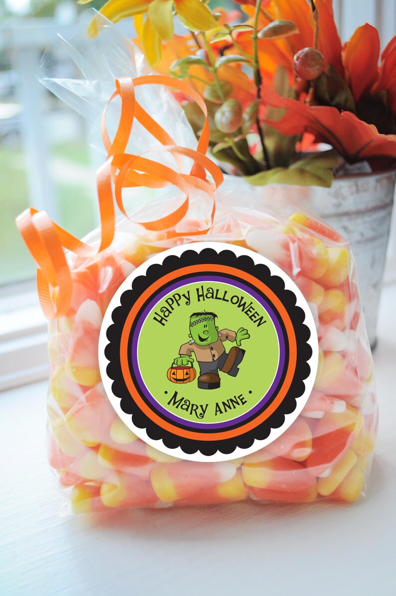May include: A round sticker with a black scalloped border, an orange and black striped border, a purple border, and a green background. The sticker says "Happy Halloween" in black text and features a cartoon green Frankenstein monster holding a jack-o-lantern. The sticker also says ". Mary Anne ." in black text.