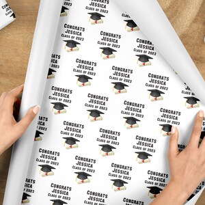 May include: White wrapping paper with a repeating pattern of graduation caps and diplomas. The text "CONGRATS JESSICA CLASS OF 2023" is printed on the paper. A roll of paper is being unrolled on a wooden surface.