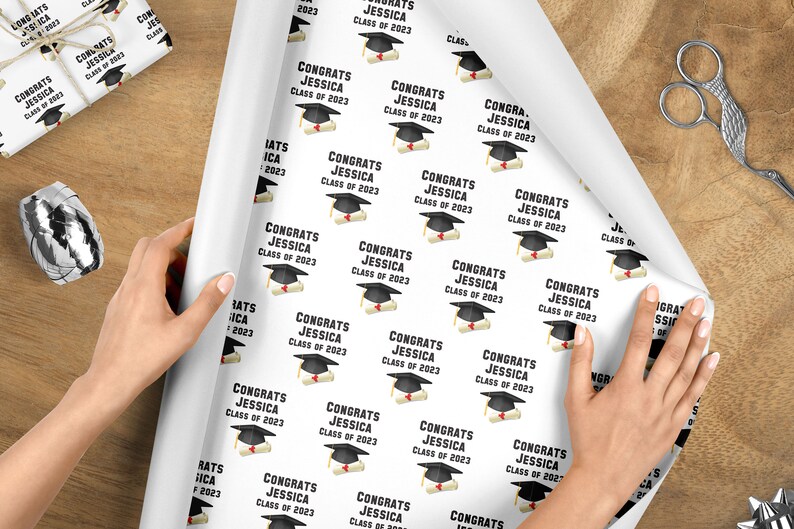 May include: White wrapping paper with a repeating pattern of graduation caps and diplomas. The text "CONGRATS JESSICA CLASS OF 2023" is printed on the paper. A roll of paper is being unrolled on a wooden surface.