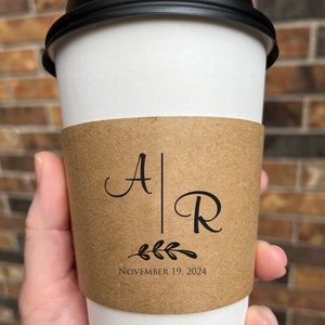 May include: A white paper coffee cup with a black lid, held by a hand. The cup has a brown sleeve with the initials "A" and "R" separated by a vertical line, with a leaf design and the date "November 19, 2024".